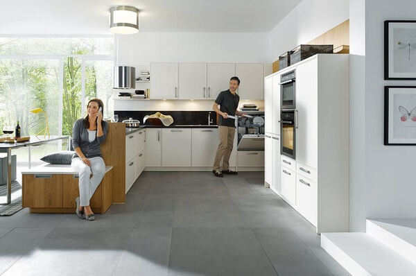 Moderne helle Einbauküche mit großen Fenstern, Kochinsel und zwei Personen beim Kochen.